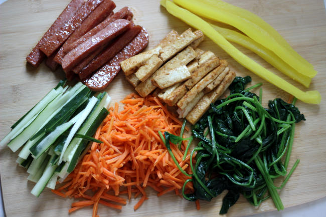 Pile of cooked shredded carrots, spinach, spam, tofu, cucumber, and pickled radish.