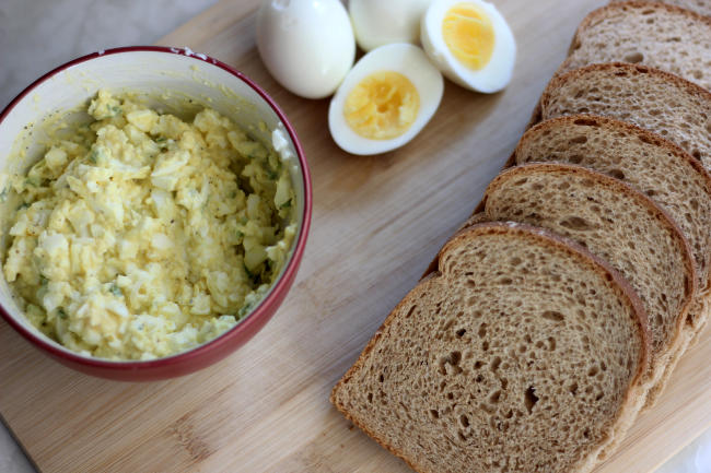 A bowl filled with egg salad, six slices of bread, and three eggs all laid out on a cutting board.